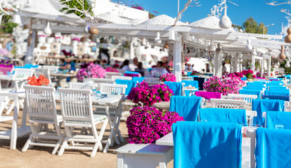 View of restaurant or cafe and bougainvillea flowers on beach in Gumusluk, Bodrum city of Turkey. Aegean seaside style colorful chairs, tables and flowers in Bodrum town near beautiful Aegean Sea.