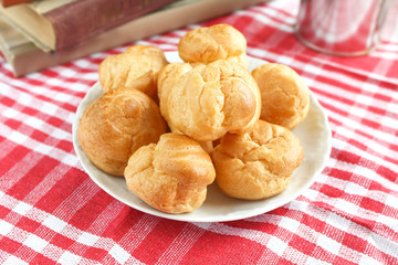 homemade small cakes profiterole choux pastry with custard next to a white cup with tea and books on a table with a rustic tablecloth in a red-white cage