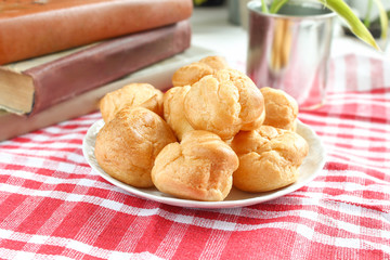 homemade small cakes profiterole choux pastry with custard next to a white cup with tea and books on a table with a rustic tablecloth in a red-white cage