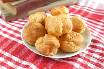homemade small cakes profiterole choux pastry with custard next to a white cup with tea and books on a table with a rustic tablecloth in a red-white cage