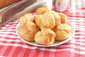 homemade small cakes profiterole choux pastry with custard next to a white cup with tea and books on a table with a rustic tablecloth in a red-white cage