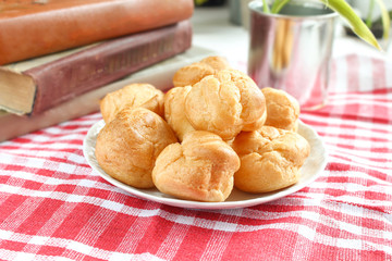 homemade small cakes profiterole choux pastry with custard next to a white cup with tea and books on a table with a rustic tablecloth in a red-white cage
