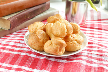 homemade small cakes profiterole choux pastry with custard next to a white cup with tea and books on a table with a rustic tablecloth in a red-white cage