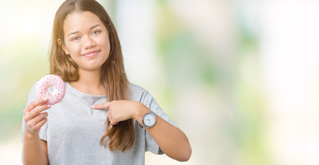 Young beautiful woman eating pink donut over isolated background with surprise face pointing finger to himself