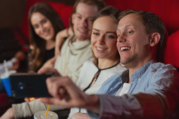 Selective focus of joyful man sitting with young friends in cinema and taking selfie on phone. Company looking at camera, laughing and posing while eating snack. Concept of fun and movie time.