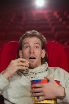Front view of amazed man with big eyes and open mouth eating tasty popcorn in cnema. Man sitting alone in hall and watching interesting new film. Concept of movie and entertainment.