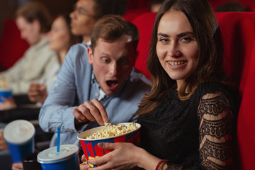 Side view of young girl looking and camera and posing while cunny man stealing popcorn at background. Cheerful friends relaxing and watching interesting movie in cinema. Concept of entertainment.
