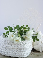 Close up crochet white basket with branch of white shrub rose,a ball of white yarn and a crochet hook on wooden table. White background. Homemade decoration, crafts and arts, hobby activity.