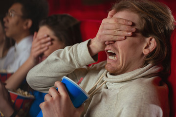 Side view of terrified man sitting in cinema and watching terrible horror. Afraid male keeping drink, closing eyes with hand and shouting in movie hall. Concept of thriller and entertainment.