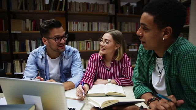 Three young students study in the school library, talking and using laptop for researching online.