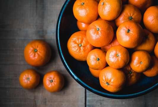Bowl of mandarin oranges on wooden table with some scattered on table.
