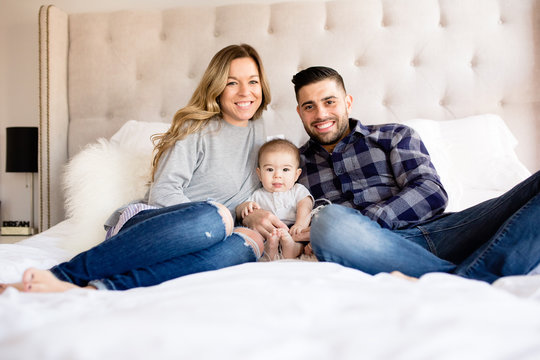 Mom Dad And Baby Sitting On Bed And Smiling At Camera