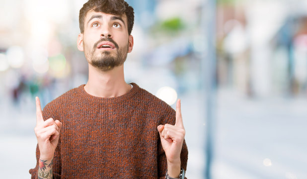Young handsome man wearing winter sweater over isolated background amazed and surprised looking up and pointing with fingers and raised arms.