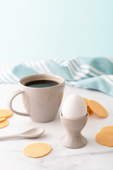 Boiled egg in ceramic egg cup, cup of coffee and thin crispy corn chips on background of beautiful white peony. Breakfast concept. Copy space.