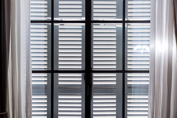 gray wooden window shutters protect the room from excessive sunlight, a window with closed shutters and open curtains of white textiles.