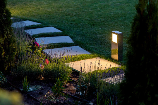 marble path of square tiles illuminated by a lantern made of metal glowing with a warm light in a backyard garden with a flower bed and a lawn.