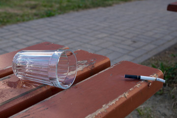 A smoking cigarette thrown on a brown bench and an emptied glass against a background of green...