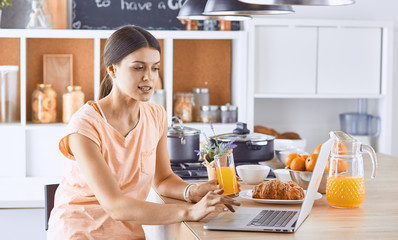 Smiling pretty woman looking at mobile phone and holding glass of orange juice while having breakfast in a kitchen