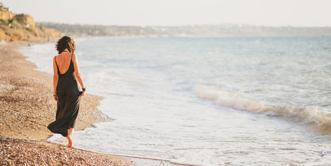 Young woman in black dress walking barefoot on beach near ocean. Summer weekend