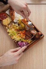 Man is eating grilled pork shish-kebab, made on coal grill, served with other bbq meat treats, coleslaw and chips on a wooden tray