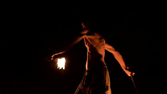 Low key. Young male with long hair and bare torso rotates burning torch outdoors on a black night video slow motion. Modern fakir does tricks with a burning staff