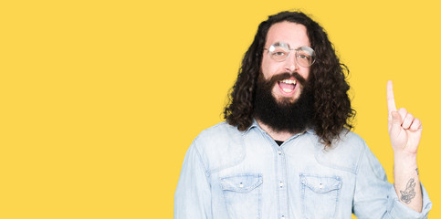 Young hipster man with long hair and beard wearing glasses showing and pointing up with finger number one while smiling confident and happy.