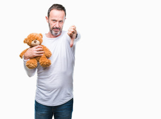 Middle age senior hoary man holding teddy bear over isolated background with angry face, negative sign showing dislike with thumbs down, rejection concept