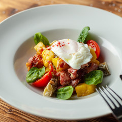 Close up of rustic salad with, tomatoes, chorizo sausage, poached egg, spinach, potato and pickled cucumber for nutritious dinner, served on wooden table