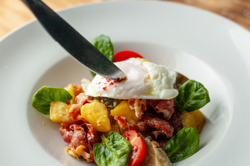 Cutting poached egg with yellow yolk on rustic salad with, tomatoes, chorizo sausage, spinach, potato and pickled cucumber served on wooden table