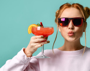 Young woman in stylish sunglasses raises her glass with tropic cocktail strawberry margarita to us and sends a kiss