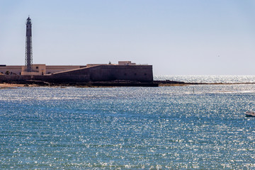 Fototapeta premium Shimmering Atlantic Ocean water surface with part of the Castle of San Sebastian and the lighthouse in Cadiz, Province of Cadiz, Andalusia, Spain seen from Playa la Caleta