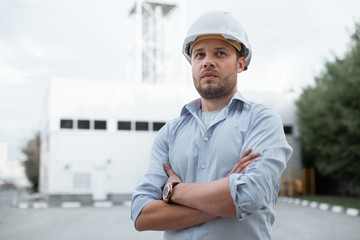 Young handsome male engineer in hardhat standing on the background of water or gas pipes. Concept of boiler station. Pipeline accessories closeup. Heating station manager doing his job outdoor. .