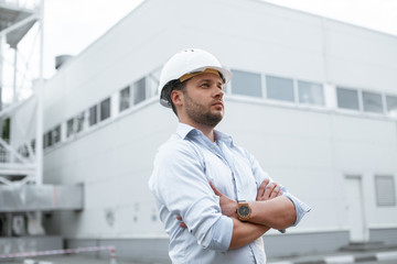 Young handsome male engineer in hardhat standing on the background of water or gas pipes. Concept of boiler station. Pipeline accessories closeup. Heating station manager doing his job outdoor. .