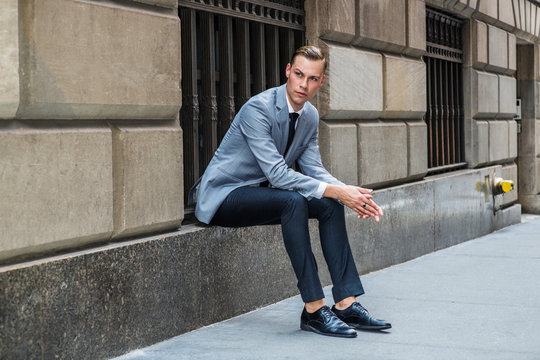 Young European Businessman traveling in New York City, wearing gray blazer, white shirt, black tie, pants, leather shoes, sitting on old street on window frame against wall, relaxing, waiting..