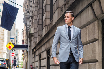 Young European Businessman traveling in New York City, wearing gray blazer, white shirt, black tie, black pants, walking on vintage street with high buildings, flag, cars, looking away..
