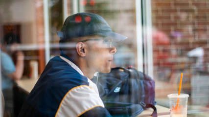 Chicago, IL/United States-June 12th 2019:  Young attractive Middle eastern male model drinks his coffee from behind the window glass at a local Chicago shop.  the student ponders about the day