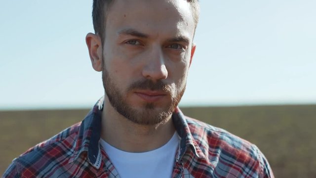 Attractive Serious Young Man Looking At Camera Standing Outdors Under Sunlight. Summer Portrait Of Handsome Farmer In Shirt In The Rural Farm Field In The Counrtyside.