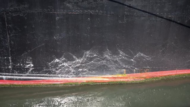 Close up detail of red plimsoll mark on large weathered black boat hull. Rippling reflections bouncing up off the water onto the boat. Slow pan from left to right.
