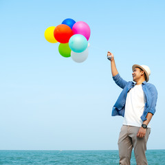 Happy Asian man holding colorful balloons against blue sky and ocean.
