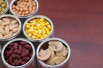 Canned green peas, beans, corn, olives and mushrooms in tin cans. Preserved food on wooden table with space for text