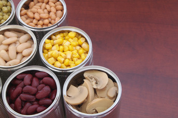Fototapeta na wymiar Canned green peas, beans, corn, olives and mushrooms in tin cans. Preserved food on wooden table with space for text