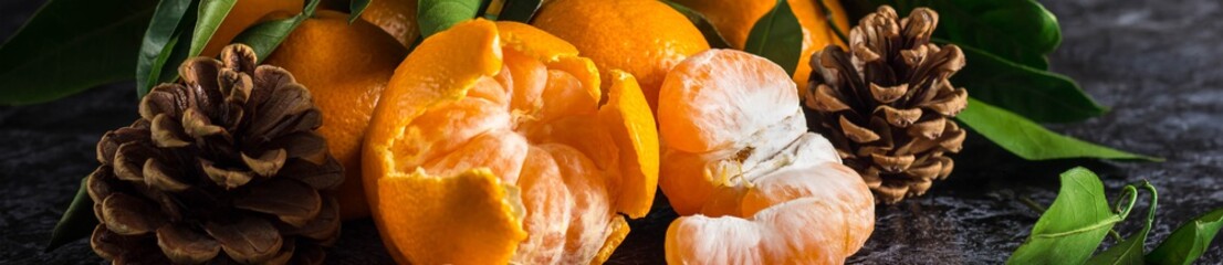 Banner of orange tangerines with green leaves on dark background. Peeled mandarin slices and cones