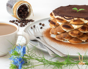 Round tiramisu, homemade no-bake traditional Italian sweet dessert. Table with cake, bowl with cocoa powder, round sponge biscuits, coffee beans and coffee with milk. White background with flowers