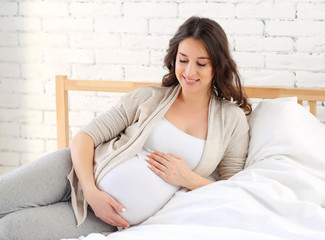 Beautiful pregnant woman with long dark hair sitting on the bed and smiling