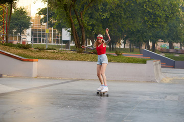 Сute blonde with tattooed arms in a red T-shirt and denim shorts with a knitted bandana on her head, in red glasses, learning to ride on a longboard in skate park, looks concentrated with hands up.