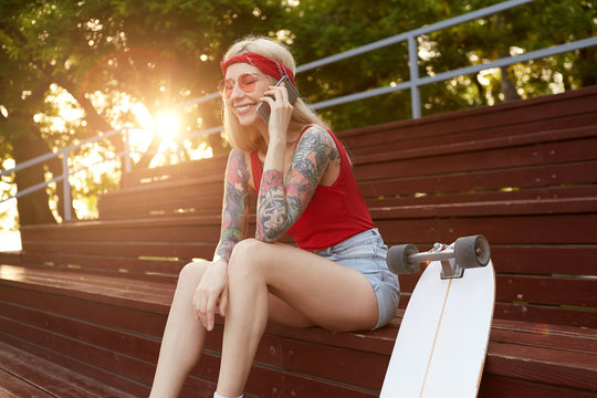Young nice laughing blonde woman with tattooed arms, talking to his friend on smartphone, hears funny joke. Wears in red T-shirt, denim shorts, with knitted bandana on her head, in red glasses.