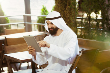 Arab saudi businessman working online with a laptop and tablet in a coffee shop or a cafe with an outdoor terrace in the background. Concept of business, finance, modern technologies, start up.
