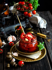 Tasty sun dried tomatoes with garlic, pepper, basil, olive oil in glass jar stands on brass plates on rustic wooden table