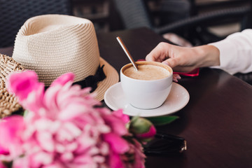 Female hands holding a cup of coffee in a cafe, on the table a bouquet of pink flowers and a summer hat