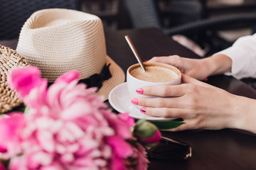 Female hands holding a cup of coffee in a cafe, on the table a bouquet of pink flowers and a summer hat